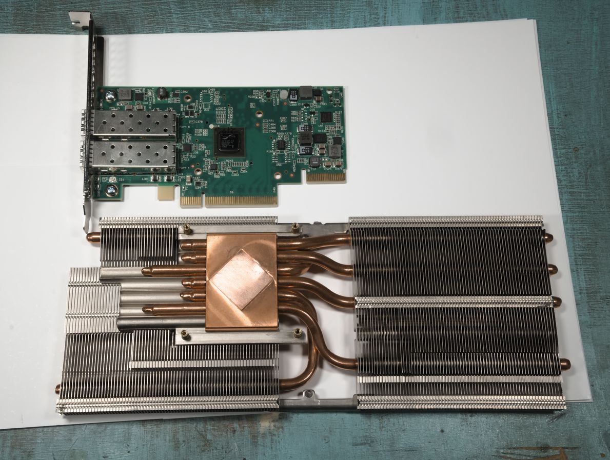 The network card with its heatsink removed. Under where the heatsink was is a small black square - the controller chip. The donor GPU heatsink is beside the card, and much larger than it. The heatsink has dense aluminium fins connected to a copper base with heat pipes.
